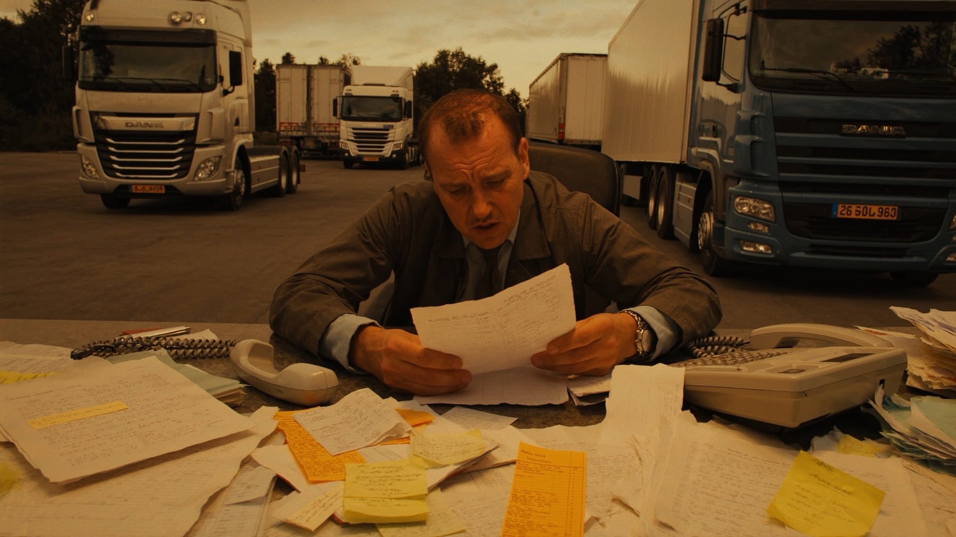 A dispatcher at a cluttered desk early in the morning, surrounded by paper transport orders and a ringing phone